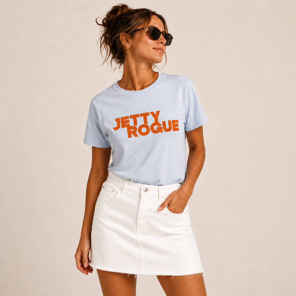 A woman modeling a light blue crewneck t-shirt with 'JETTY ROGUE' printed in orange across the chest, paired with a white denim mini skirt against a solid beige background.