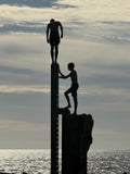 Against a dramatic sky three black figures in silhouette all try to get to the top of the same pylon at Glenelg jetty Adelaide. One stands on top, another stands below him, while a third ominous figure crawls up behind them.