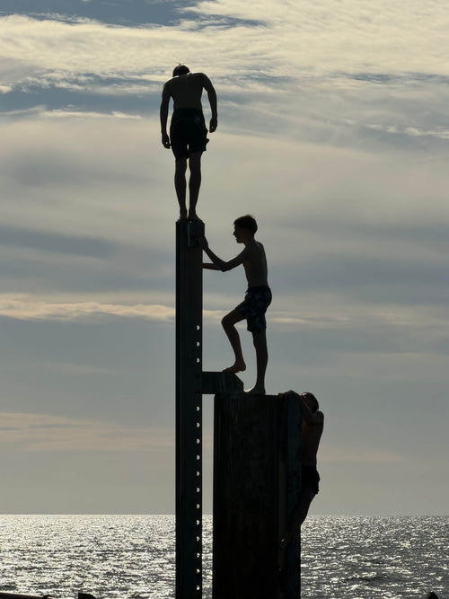 Against a dramatic sky three black figures in silhouette all try to get to the top of the same pylon at Glenelg jetty Adelaide. One stands on top, another stands below him, while a third ominous figure crawls up behind them.