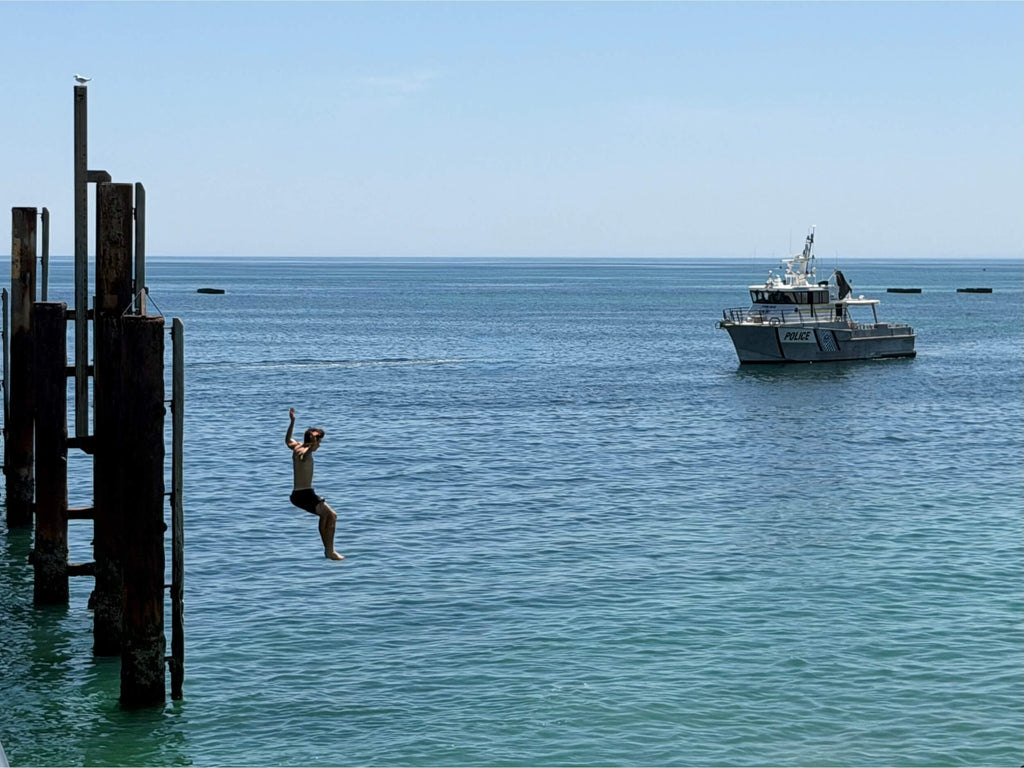 Boy jumping midair off Glenelg jetty Adelaide against a bright blue sky, with his hands in the air, directly in front of a Police boat.