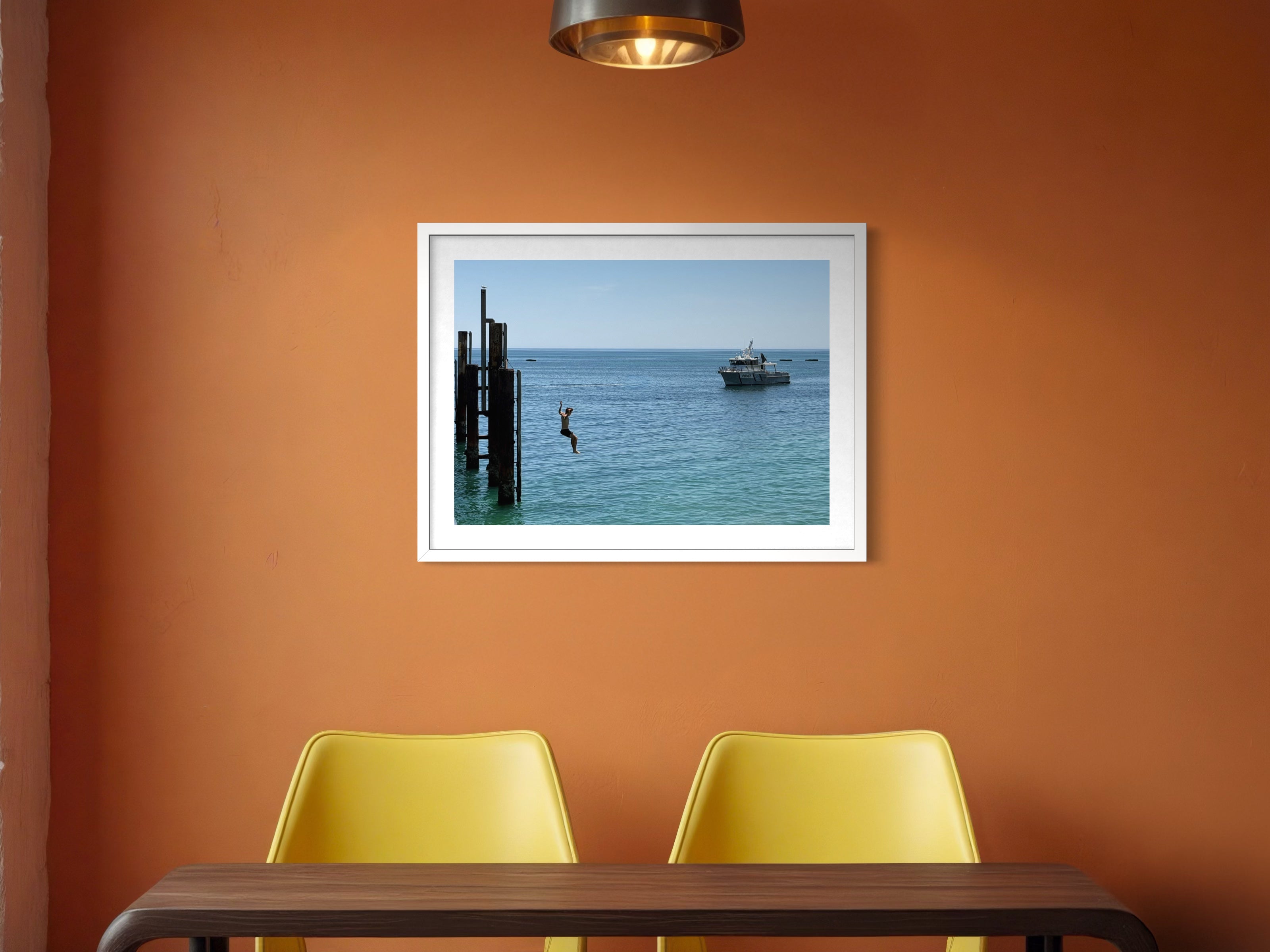 Framed picture of a dock scene on an orange wall above two yellow chairs.