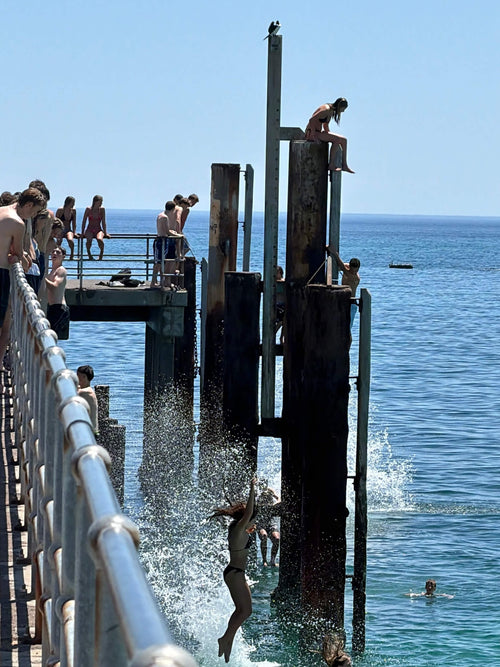 Young people at Glenelg jetty leaping and splashing into the water from pylons of different heights on a glorious Summer day with a bright blue sky and sea. 