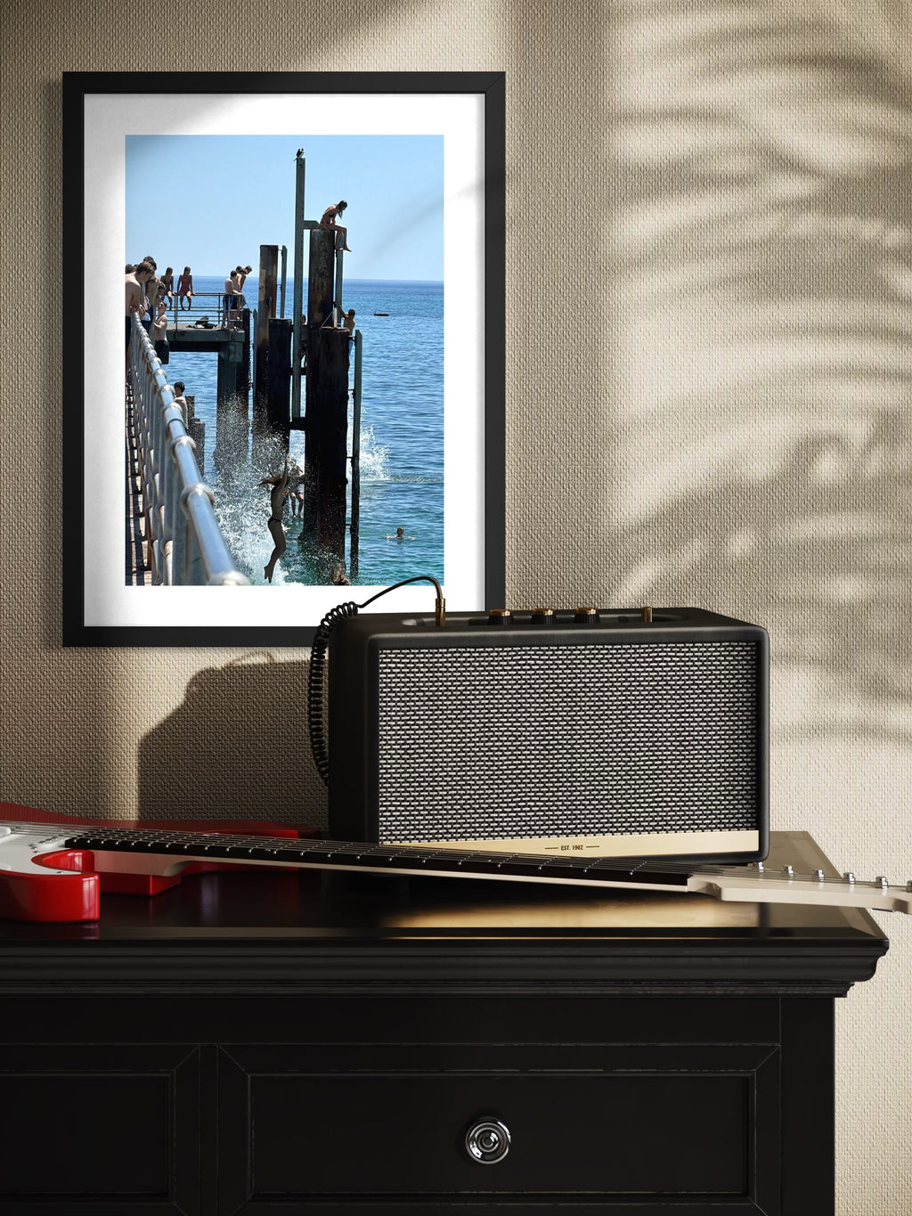 Framed photograph of a dock scene on a wall above a guitar and amplifier.