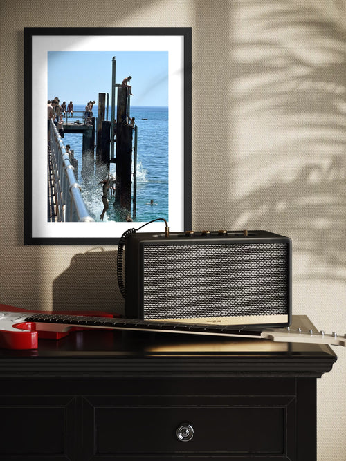 Framed photograph of a dock scene on a wall above a guitar and amplifier.