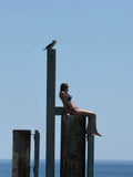 Beautiful girl resting on a pylon at Glenelg Jetty Adelaide, while a seabird rests on a pylon above her. They are both silhouetted looking the same way out to a vibrant blue sky and sea.