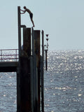 Man perched on his toes on a high pylon at Glenelg jetty South Australia. Silhouetted against a grey blue sky and sparking sea he appears hesitant to jump. 