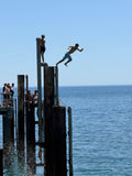 Multiple pylons at Glenelg jetty are silhouetted against a bright blue sky and sea, as a figure takes the first step off into the air.