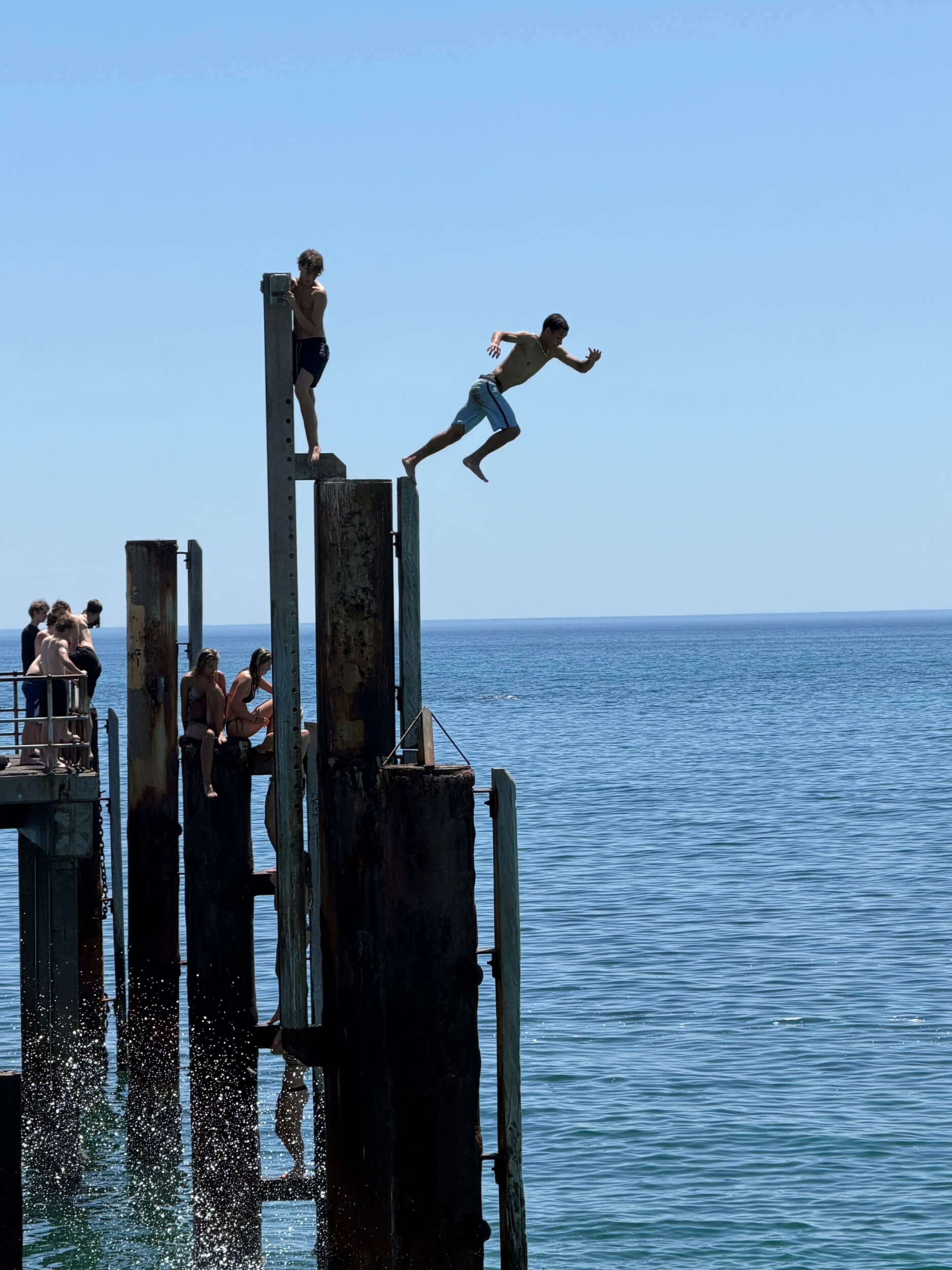 Multiple pylons at Glenelg jetty are silhouetted against a bright blue sky and sea, as a figure takes the first step off into the air.