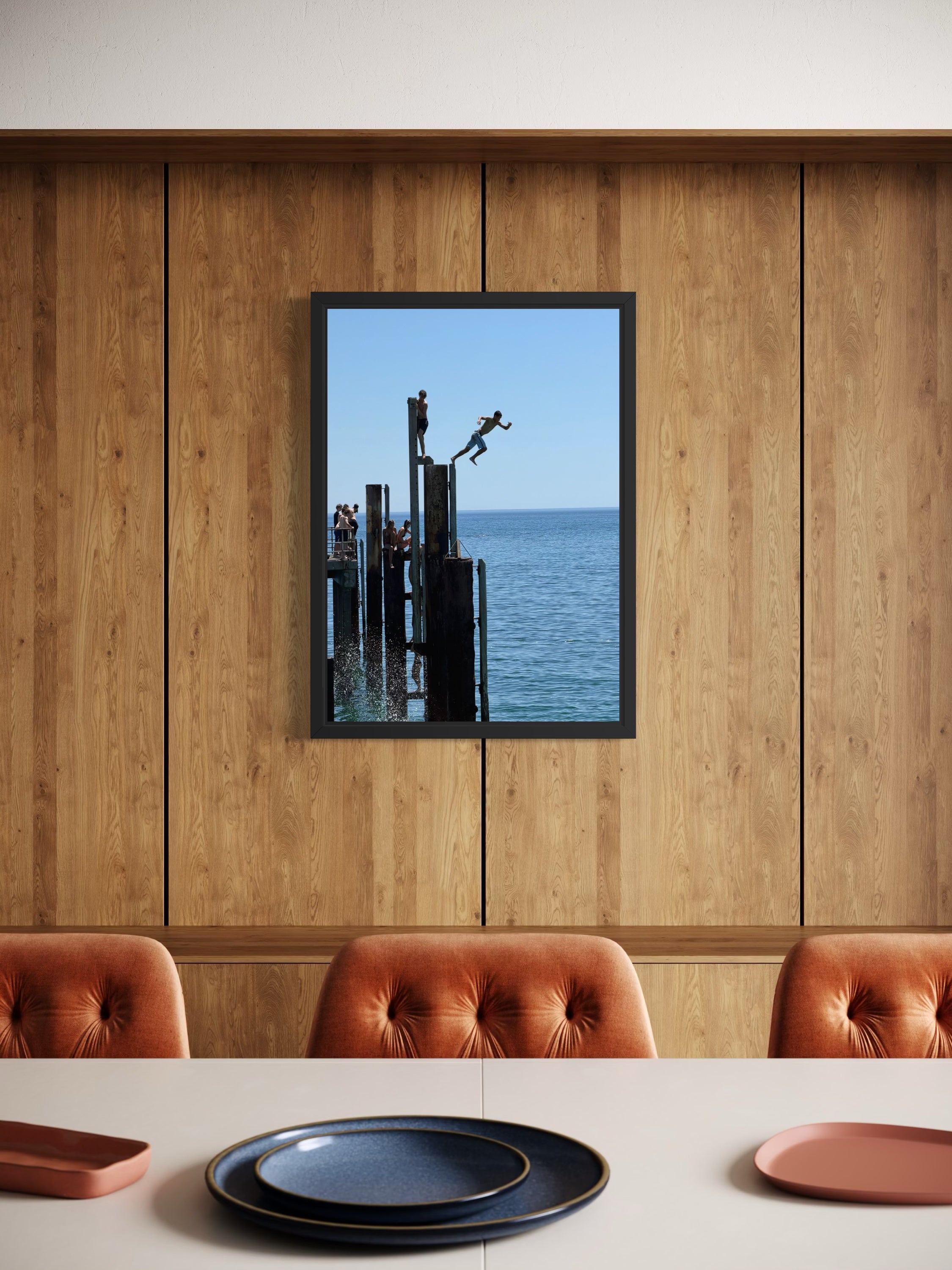 Dining area with a wooden wall, framed picture of people by water, and brown chairs.