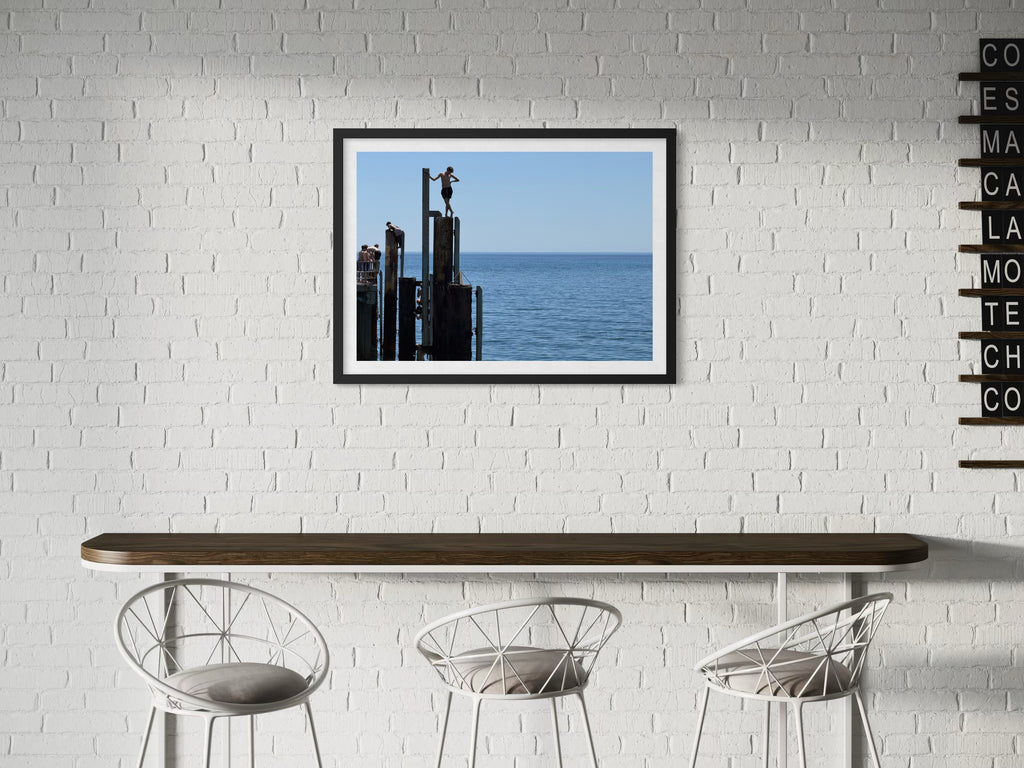 Framed artwork of a cityscape with water on a white brick wall above a table and chairs.