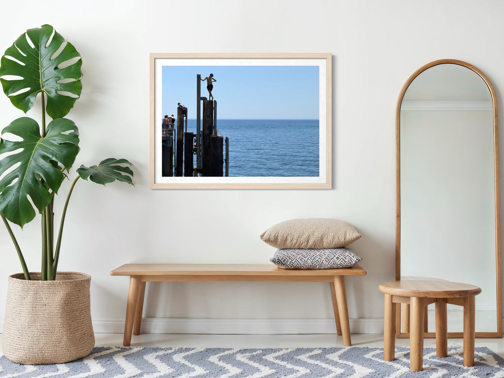 Framed artwork on a wall above a wooden bench with decorative pillows, next to a plant and mirror.