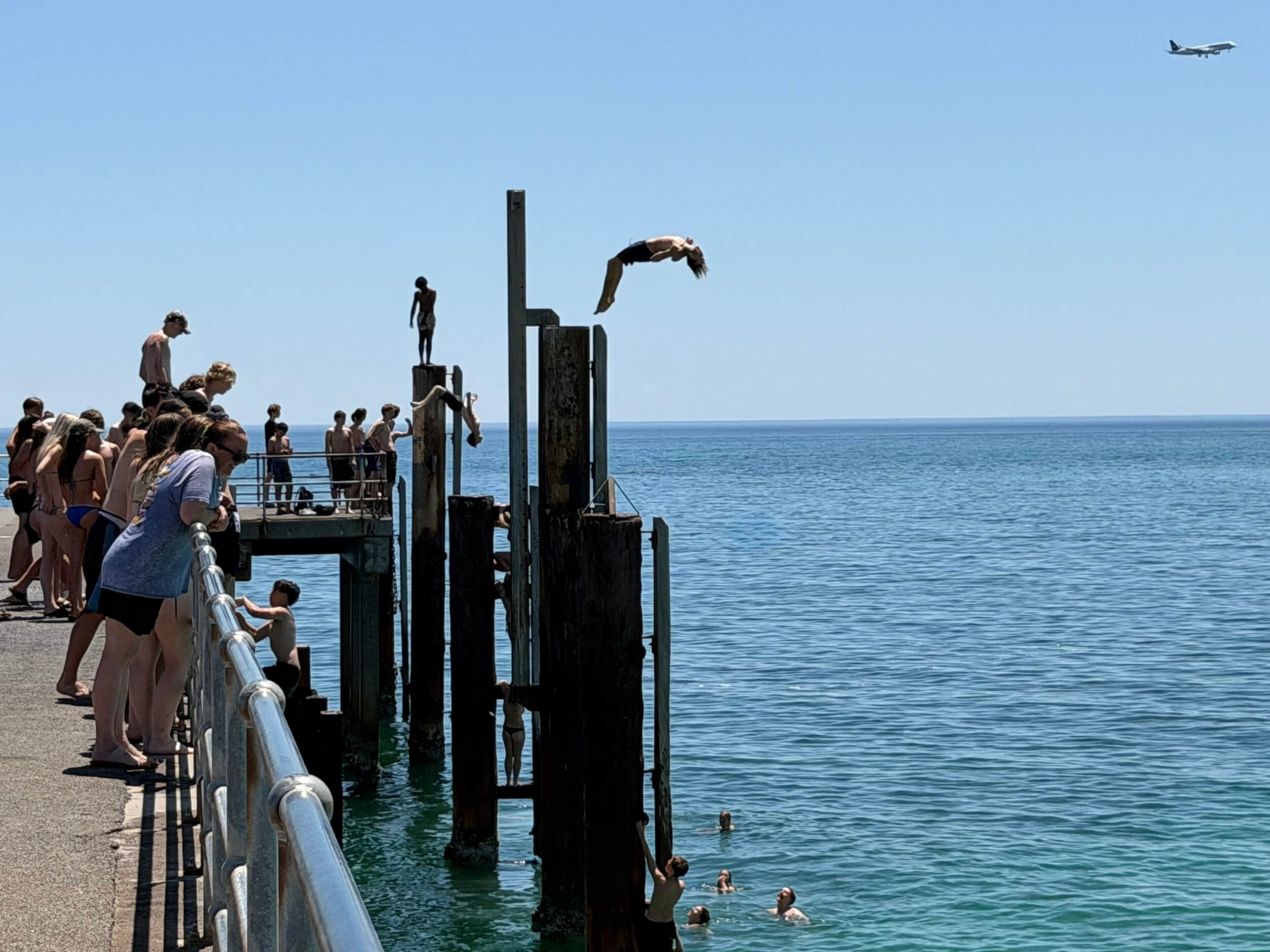Multiple figures in silhouette doing backflips and climbing up high, black pylons against a vibrant, blue sea and sky at Glenelg Jetty Adelaide. Crowd watches as a plane flies overhead.
