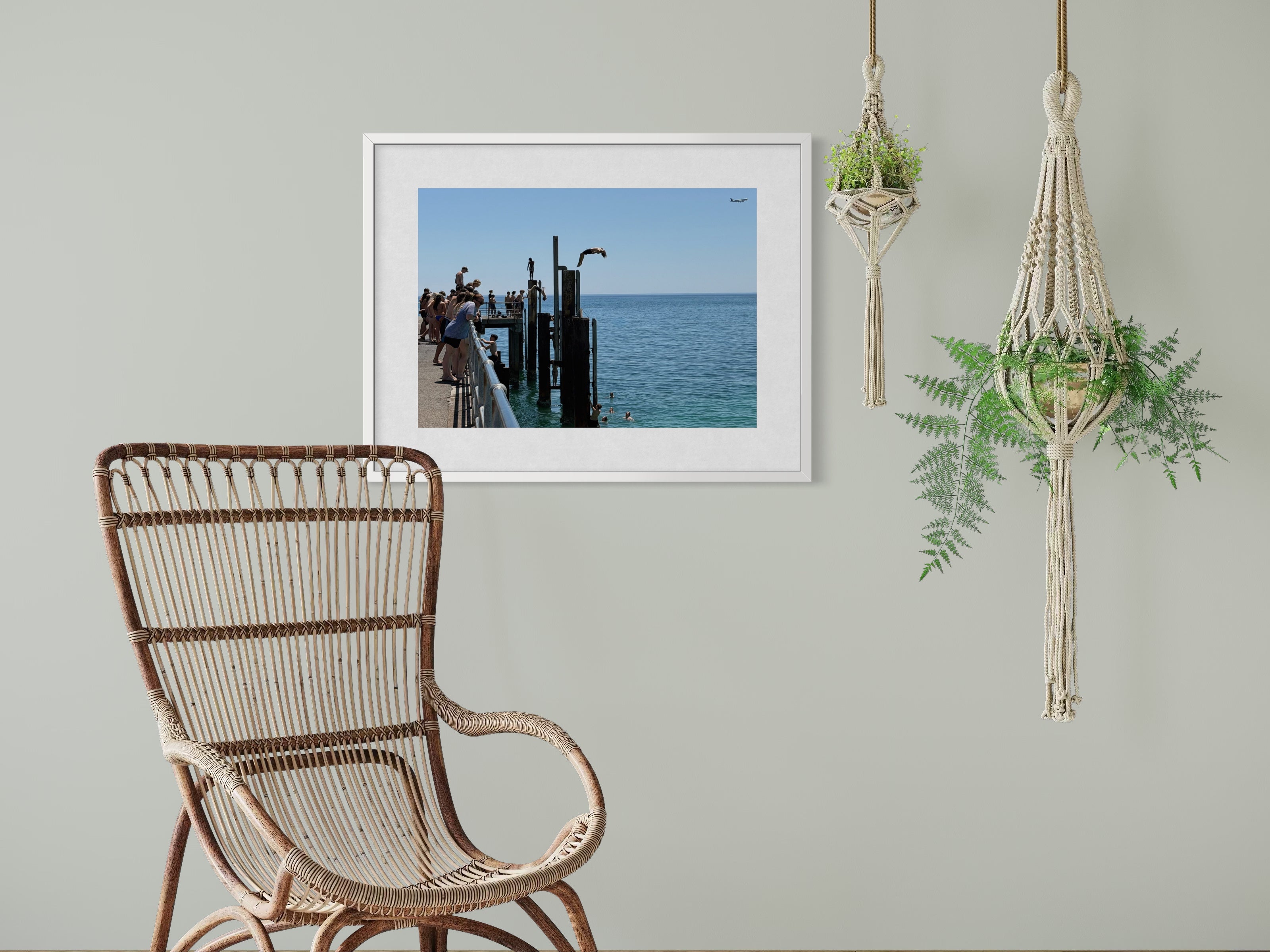 Wicker chair in a room with a framed picture of a dock, a macrame plant holder, and hanging plants.