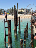 A boy stands on a tall, crusty pylon on a beautiful Summer day at Glenelg Adelaide. Aqua blue water is around him and a ferris wheel is in the background.